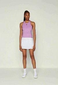 Athletische Frau steht und trägt ein ärmelloses lila Oberteil, einen weißen, plissierten Tennisrock, weiße Socken und weiße Turnschuhe vor einem schlichten Hintergrund.