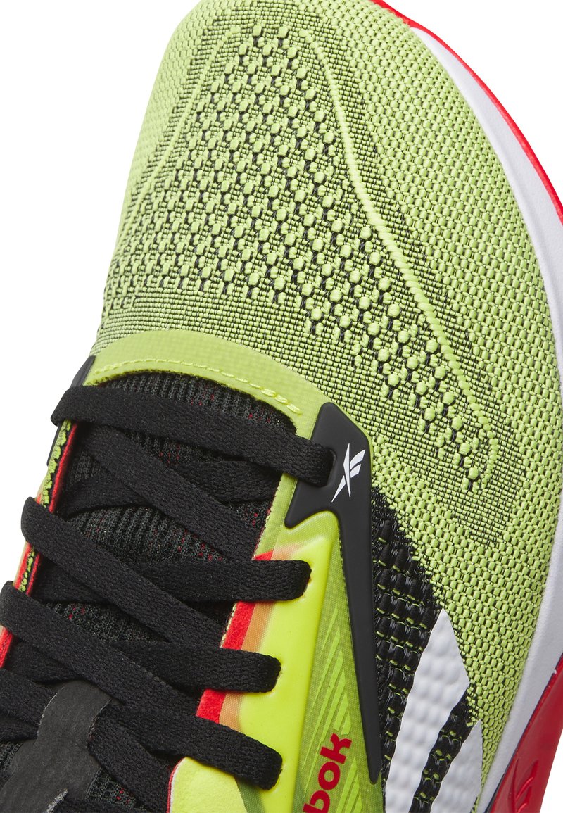 Zapatilla deportiva verde neón con parte superior texturizada, cordones negros, acentos en negro y rojo, y detalle del logo en el lateral. Diseño ligero.