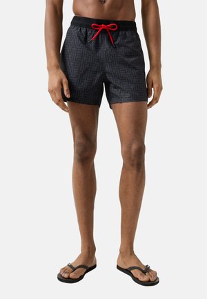 Mand iført sorte mønstrede badeshorts med rød snor og sorte klipklappere, stående mod en ensartet lys baggrund.