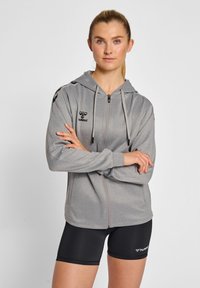 Giacca a zip grigia con cappuccio, dal morbido texture lucido, con dettagli neri e un logo sul petto, abbinata a pantaloni corti neri aderenti.