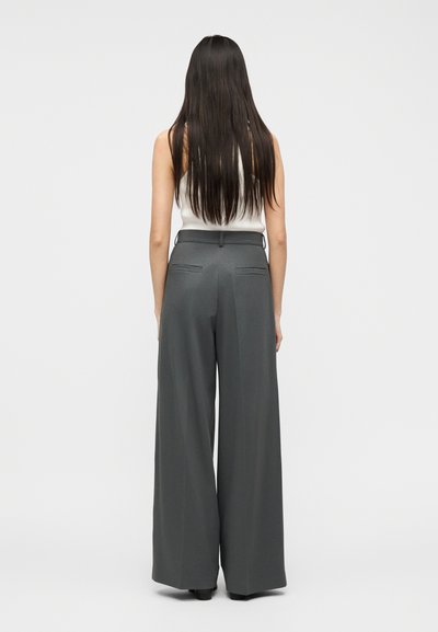 Γυναίκα με μακριά σκούρα μαλλιά, ντυμένη με λευκό αμάνικο τοπ και γκρι παντελόνι ψηλόμεσο με φαρδιά μπατζάκια, στέκεται μπροστά σε απλό φόντο.