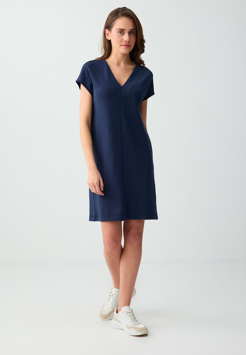 Femme debout en robe bleu marine à manches courtes et col en V avec des baskets blanches sur un fond gris clair uni.