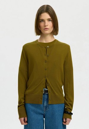Kvinde med kort brunt hår, iført olivengrøn knappet cardigan og blå jeans, stående mod en ensfarvet lys baggrund.
