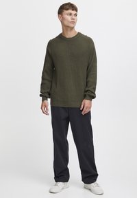 Pulover tricotat cu nervuri, de culoare verde olive, cu guler rotund, mâneci lungi și o croială lejeră, asortat cu pantaloni largi, de culoare închisă și adidași albi.