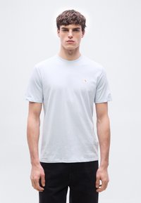 Νέος άνδρας με κοντά σγουρά μαλλιά, ντυμένος με ανοιχτό μπλε μπλουζάκι Calvin Klein και μαύρο παντελόνι, σε απλό ανοιχτό φόντο.