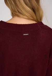 Pull en bourgogne avec une texture douce, un col large et des bordures côtelées, arborant une petite étiquette en métal au dos.