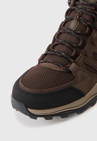 Brąsowe buty trekkingowe z teksturowaną siateczkową cholewką, zamszowymi akcentami, czarnym gumowym noskiem i podeszwą zaprojektowaną pod kątem przyczepności. Posiadają czarne i pomarańczowe sznurowadła.