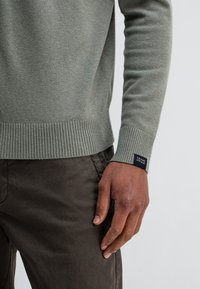 Lichtgroene gebreide trui met ribbels aan de manchetten en onderkant. Bevat een label op de manchette. Gepaard met donkerbruine broek.