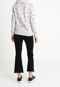 Grauer Pullover mit rotem Lippenabdruck, Schlauchhals, gekrempelten Ärmeln, kombiniert mit schwarzen ausgestellten Jeans und schwarzen Turnschuhen mit weißen Sohlen.