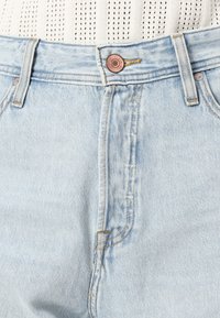 Jeans de mezclilla azul claro con un botón de cobre y costuras amarillas. La tela tiene un acabado ligeramente texturizado con un diseño clásico de cinco bolsillos.