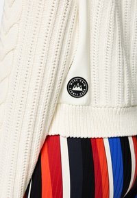 Weißer Strickcardigan mit einem Zopfmuster, einem gerippten Saum und einem runden schwarzen LOGO-Patch auf einem kontrastierenden Stoff. Mehrfarbiges Streifenmuster darunter.