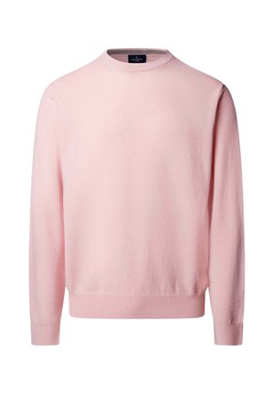 Joven de pie con un jersey rosa claro, pantalones marrón oscuro con un cinturón negro y zapatos de vestir negros, frente a un fondo gris liso.