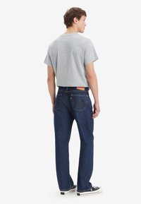 Homme portant un t-shirt gris, un jean droit bleu foncé et des baskets basses noires, debout et regardant sur le côté devant un fond blanc.