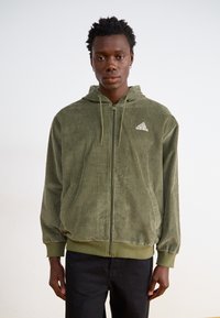 Olīvzaļš korodura mētelis ar rāvējslēdzēju, kapuci, priekšējām kabatām un mazu baltu Adidas logo uz krūts. Iezīmēts ar ribotām piedurknēm un apakšmalu.