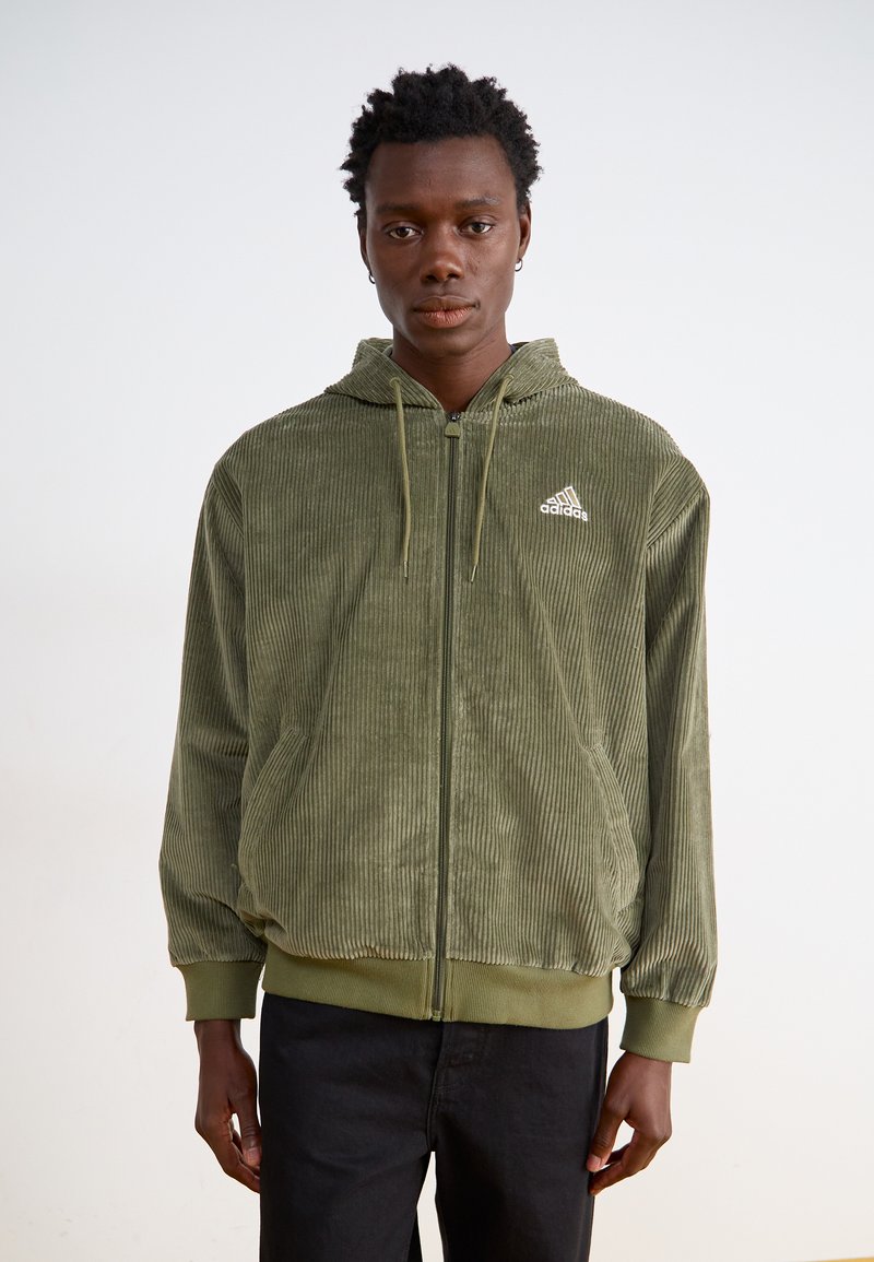 Olīvzaļš korodura mētelis ar rāvējslēdzēju, kapuci, priekšējām kabatām un mazu baltu Adidas logo uz krūts. Iezīmēts ar ribotām piedurknēm un apakšmalu.