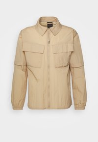 Giacca leggera beige con zip frontale, ampie tasche sul petto e colletto. Presenta maniche leggermente arricciate e una texture liscia.