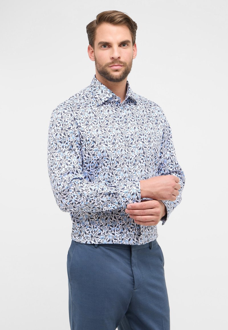 Camicia a maniche lunghe di colore bianco con motivo floreale blu, dotata di colletto aperto e polsini con bottone. Indossata con pantaloni blu.