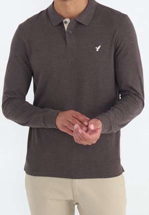 Man draagt een donkergrijs poloshirt met lange mouwen en een wit vogel-logo op de borst, beige broek, en houdt zijn handen voor zich vast.