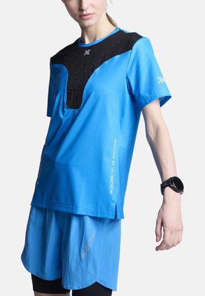 Vrouw draagt een blauw-zwart sportshirt met korte mouwen en bijpassende blauwe shorts, met een zwarte smartwatch om haar linkerpols.