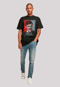 Black cotton t-shirt featuring a colorful graphic of a man’s face, text "HILL" below, paired with distressed blue jeans and white sneakers.