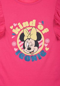 Camiseta rosa con mangas acampanadas que presenta un gráfico de un personaje de dibujos animados con un lazo amarillo, rodeado de texto decorativo y corazones.