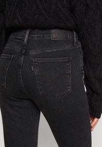 Pantalones vaqueros de mezclilla negra con una forma ajustada. Presenta dos bolsillos traseros con un patrón de costura distintivo y una etiqueta en la cintura.