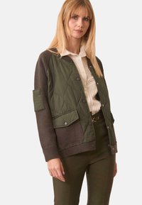 Mujer vistiendo una chaqueta acolchada y de punto en verde oliva sobre una blusa blanca, combinada con pantalones de tiro alto en verde oliva y un cinturón marrón.