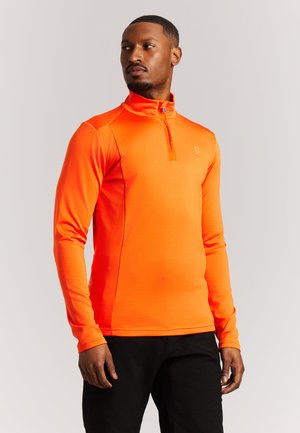 Mann trägt ein leuchtend orangefarbenes Langarm-Quarter-Zip-Oberteil und schwarze Hose, steht und schaut seitlich vor einfarbigem Hintergrund.
