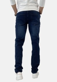 Jeans in denim blu scuro con vestibilità slim, dotati di due tasche posteriori, una texture liscia e una leggera elasticità. Indossati con sneakers bianche.
