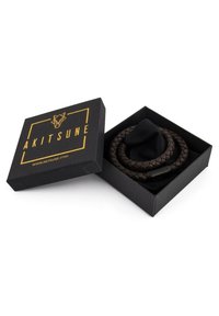 Pulsera de cuero trenzado marrón en una caja negra con marca dorada. Forro de tela suave dentro de la caja. Diseño minimalista.