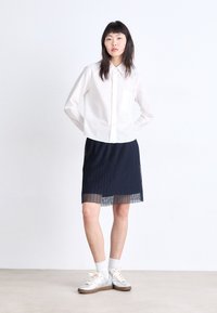 Camisa de rayas blancas de manga larga con un bolsillo en el pecho, combinada con una falda azul marino con pliegues y zapatillas plateadas sobre un fondo neutro.