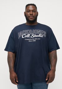 Navy blue short-sleeve t-shirt with a round neckline, featuring white text branding “JACK & JONES Coll. Studio” on the front.
