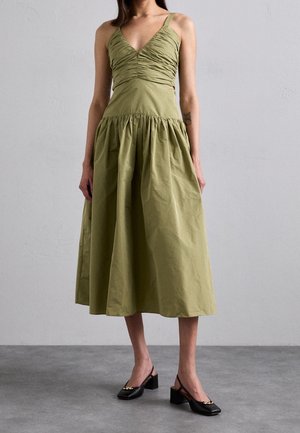 Kvinde iført en olivengrøn, ærmeløs ruched midi-kjole med en tætsiddende overdel og en udvidet nederdel, stylet med sorte blokhælede slingback-sko.