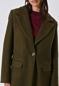 Cappotto verde oliva, strutturato, con strisce verticali, ampie rever, chiusura a un bottone e due tasche frontali. Indossato sopra un maglione bordeaux.