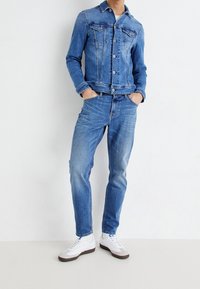 Blå denimjacka med knappstängning och två bröstfickor, kombinerad med ljusblå jeans och vita sneakers. Slät textur, avslappnad design.