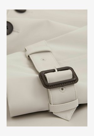 Tkanina svetložute trench jakne s tamnimi gumbi in sponko z rjavimi akcenti. Ima gladko teksturo in kontrastne šive.