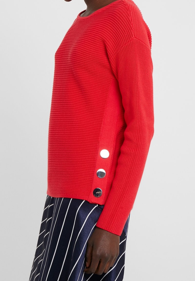 Personne portant un pull rouge texturé avec trois gros boutons argentés sur le côté, associé à une jupe bleu marine à rayures verticales blanches.