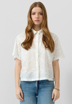 Junge Frau mit langen, hellbraunen Haaren, die ein weißes, kurzärmeliges, besticktes Hemd mit Knöpfen und blaue Jeans vor einem schlichten Hintergrund trägt.