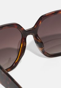 Lunettes de soleil en écaille de tortue avec des verres bruns dégradés, présentant une monture géométrique audacieuse et une texture lisse, mettant en valeur le motif unique.