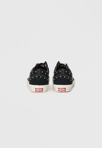 Adidași din suede negru, cu ținte argintii, tălpi din cauciuc alb și un logo roșu pe călcâi. Vedere din spate care arată un design de tip low-top.