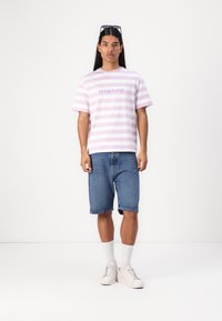 Rutet lilla og hvite t-skjorte med "GUESS JEANS"-logo, kombinert med denimshorts og hvite joggesko, brukt med hvite crew-sokker.