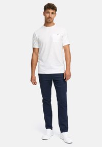 Mand iført en hvid t-shirt med korte ærmer og lomme, marineblå slim-fit bukser og hvide sneakers, stående foran en ensfarvet lys baggrund.
