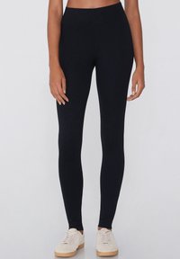 Leggings noirs en tissu extensible, avec une taille haute. Portés avec des baskets blanches sur un fond neutre.