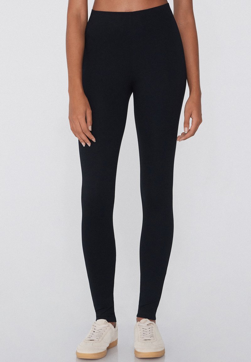 Leggings noirs en tissu extensible, avec une taille haute. Portés avec des baskets blanches sur un fond neutre.