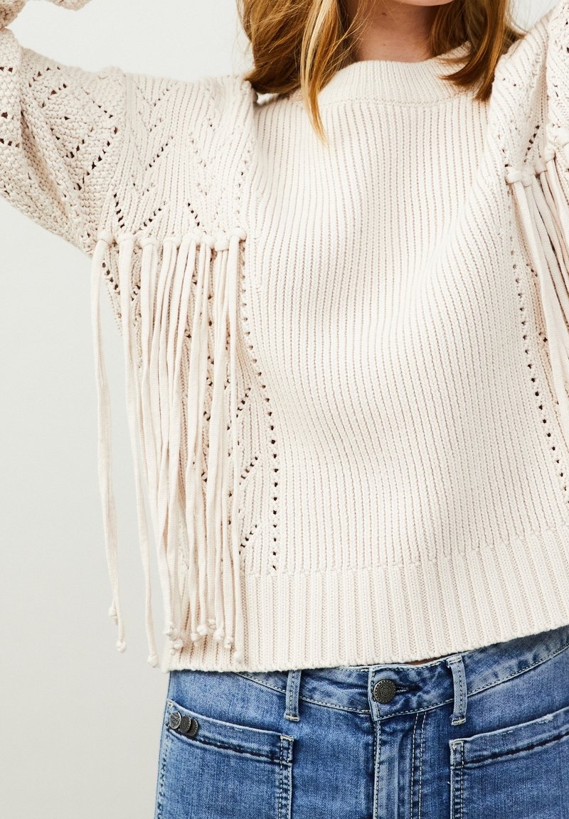 Maglione beige lavorato a maglia con texture a costine, contrassegnato da dettagli in frange sulle maniche e un collo rotondo, abbinato a jeans in denim chiaro.