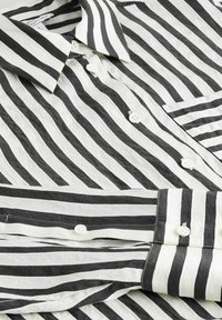 Chemise à rayures noires et blanches avec boutons. Tissu léger, col classique et poche avant. Boutons blancs et texture lisse.