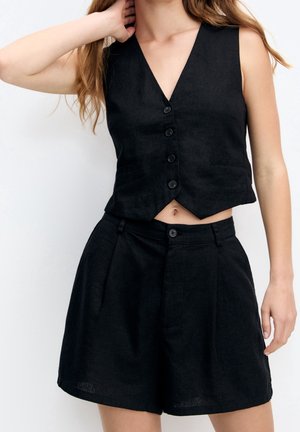 Zwarte linnen vest met een V-hals en vijf knopen, gecombineerd met zwarte linnen shorts met hoge taille, voorzien van plooien en knoopsluiting.