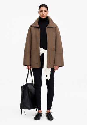 Femme portant une veste marron oversize, un col roulé noir, des leggings noirs, des chaussures noires, tenant un grand sac cabas noir avec un pull blanc noué à la taille.