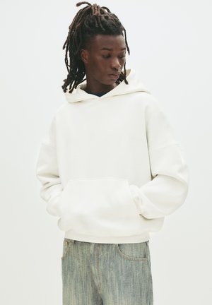 White hoodie with a large front pocket and drawstring hood, featuring a relaxed fit and ribbed cuffs. Paired with light denim jeans.