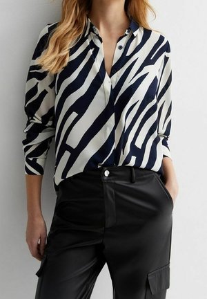 Femme portant un chemisier boutonné à rayures zébrées noir et blanc avec un pantalon noir taille haute, une main dans la poche, devant un mur uni.